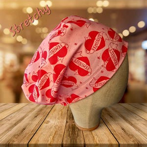 May include: A pink, stretchy cap with a repeating pattern of red hearts and the word "FEMINIST" on a banner. The word "Stretchy" is written in pink script across the top left of the cap.