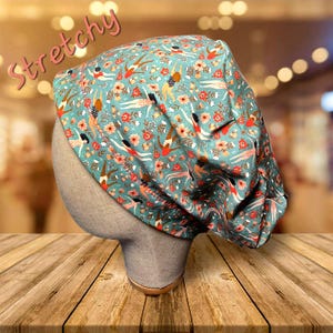 May include: A teal surgical scrub cap with a pattern of swimmers and flowers. The cap is on a mannequin head and has the word "Stretchy" in pink script across the top. The cap is made of a soft, flexible material.
