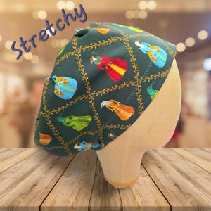 May include: Dark green, stretchy hat with a repeating pattern of colorful figures and gold-colored vines. The word "Stretchy" is written in blue script across the top. The hat is displayed on a mannequin head.
