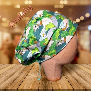 May include: A green and blue patterned scrub cap with a landscape design featuring trees, houses, and birds. The cap has a drawstring and the word "Stretchy" in pink. The cap is displayed on a mannequin head.