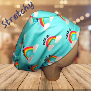 May include: A turquoise, stretchy cap featuring a repeating pattern of rainbows and clouds. The text "OVER it." is printed near the rainbows. The word "Stretchy" is written in a cursive font in the upper left corner.