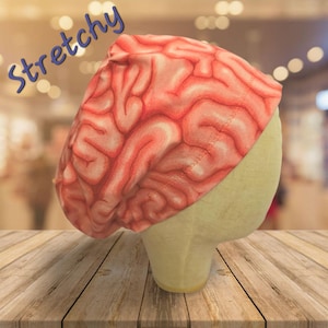 May include: A pink and red brain-patterned scrub cap is displayed on a mannequin head. The word "Stretchy" is written in blue cursive across the top left of the image. The cap is made of a soft, flexible material.