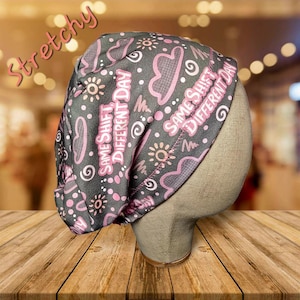 May include: A gray and pink patterned scrub cap with the text "Same Shift Different Day" in pink. The cap features a repeating pattern of clouds, suns, and dots. The word "Stretchy" is written in pink script across the top.
