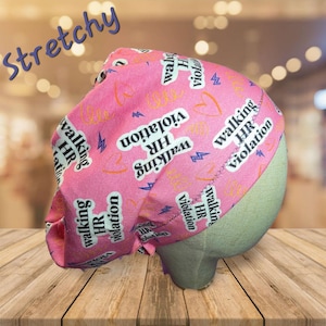 May include: Pink scrub cap with the text "Walking HR Violation" in white, along with lightning bolts and hearts. The word "Stretchy" is written in blue script. The cap is displayed on a mannequin head.