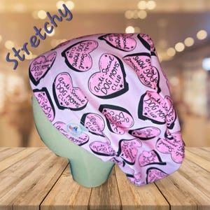 May include: A pink and white patterned scrub cap with the text "Anti-Social Dog Moms Club" in black. The cap is made of a stretchy material and features heart-shaped designs. The word "Stretchy" is written in blue.