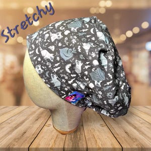 May include: A dark gray, stretchy scrub cap with a whimsical pattern of raccoons, trees, moons, and snowflakes. The word "Stretchy" is written in blue script across the top. A small, colorful tag is sewn on the side.