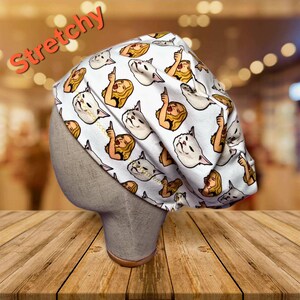 May include: A white, stretchy cap featuring a repeating pattern of cartoon cats and women pointing upwards. The word "Stretchy" is written in orange script across the top. The cap is displayed on a mannequin head.
