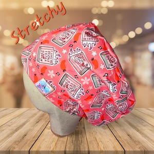 May include: A pink surgical scrub cap with a repeating pattern of books, chili peppers, and flowers. The text "I like my books extra spicy" is printed on the books. The word "Stretchy" is written across the top.