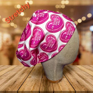 May include: A white and pink patterned cap with heart-shaped candy graphics. Each candy heart has the text "I'M LITERALLY JUST A GIRL". The word "Stretchy" is written in orange across the top of the image.