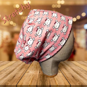 May include: Pink skull cap with a repeating pattern of white skulls with red bows. The word "Stretchy" is written in pink script across the top left of the image. The cap is displayed on a mannequin head.