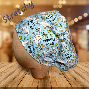May include: A light blue surgical scrub cap with a whimsical pattern of white geese, daisies, and hearts. The words "Silly Goose!" are scattered across the fabric. The word "Stretchy" is written in a script font.