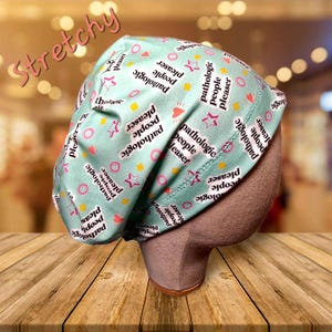 May include: A light green, stretchy surgical scrub cap with a repeating pattern of the words "pathologic people pleaser", stars, hearts, and circles. The word "Stretchy" is written in pink script across the top.
