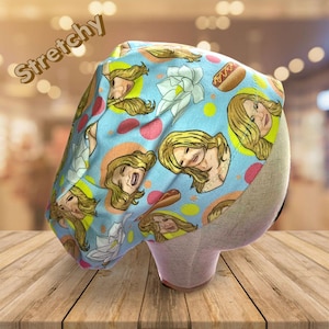 May include: A light blue, stretchy cap with a repeating pattern of blonde women's faces, hot dogs, and white bows. The cap is decorated with pink and yellow polka dots. The word "Stretchy" is printed in a cursive font across the top of the cap.