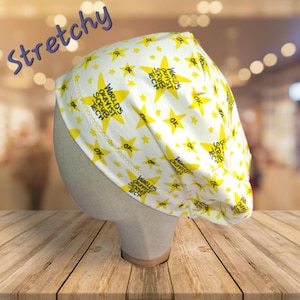May include: A white and yellow scrub cap with a star pattern and the text "WORLD'S OKAYIST SCRUB TECH". The word "Stretchy" is written in blue on the top left of the cap.