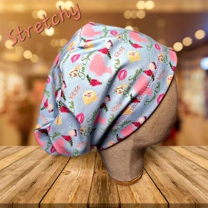 May include: A light blue, stretchy cap with a repeating pattern of pink lips, roses, teacups, and envelopes. The words "Tea Spiller" and "Social Club" are also printed on the cap. The word "Stretchy" is written in pink cursive.