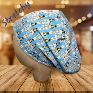 May include: A blue and white patterned scrub cap with a repeating design of figures. The word "Stretchy" is written in blue script across the top. The cap is displayed on a mannequin head.