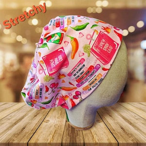 May include: A pink, stretchy scrub cap with a repeating pattern of chili peppers, books, and text. The text includes phrases like "Spicy Book Club" and "Friends to Lovers." The word "Stretchy" is printed in orange across the top.