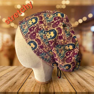 May include: A patterned scrub cap featuring a repeating design of a woman's face, crosses, and bottles in burgundy, teal, and cream colors. The word "stretchy" is written in orange across the top. The cap is on a mannequin head.