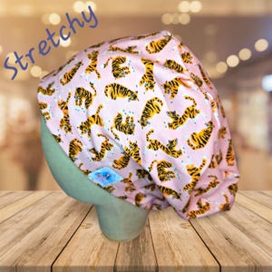 May include: Pink scrub hat with a tiger print. The hat is stretchy and has a tie at the back.