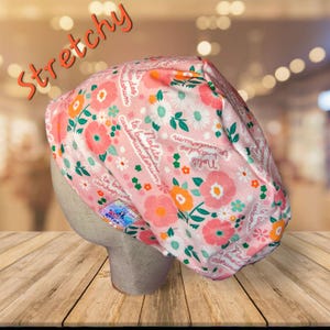 May include: A pink floral patterned scrub cap with the word "Stretchy" in orange script. The cap features a repeating pattern of flowers in shades of pink, orange, and white, with green leaves and stems. The cap is displayed on a mannequin head.