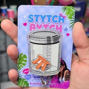 May include: An acrylic badge reel featuring a clear container graphic with a measuring scale and a small orange stool inside. The backing card displays the text STYTCH BYTCH, SASSY, PETTY, and BADGE, with a light blue checkered pattern and green leaves.