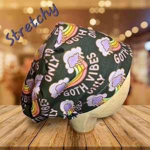 May include: A black cap with a repeating pattern of rainbows, clouds, and the words "Goth Vibes Only." The word "Stretchy" is written in blue script across the top. The cap is displayed on a mannequin head.