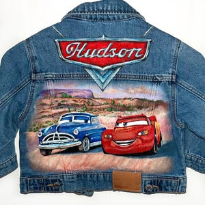 May include: A blue denim jacket featuring a painting of two cartoon cars and the word "Hudson". The artwork is on the back of the jacket, which has a brown leather patch.