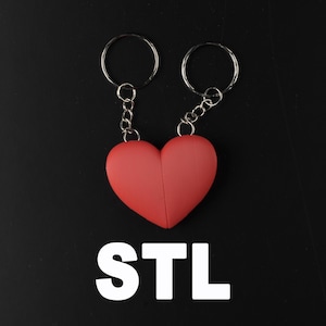 Op de afbeelding: Twee zilveren sleutelhangers die aan elkaar zijn verbonden met een rode hartvormige hanger. De hanger is met zilveren kettingen aan de sleutelhangers bevestigd. De sleutelhangers bevinden zich op een zwarte achtergrond. De tekst "STL" is in wit onder de hanger geschreven.