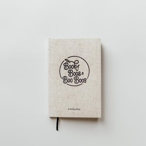 Puede incluir: Un cuaderno de textura de lino beige con un marcapáginas de cinta negra. La portada presenta el texto negro "The Book of Boos & Boo Boos" dentro de un círculo, con las palabras "A dating diary" impresas en una fuente negra más pequeña.