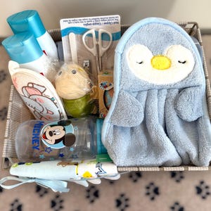 May include: A baby gift basket containing baby essentials. It features a blue penguin washcloth, baby bottles, lotion, nail clippers, a rattle, and a soft toy. The basket is beige.