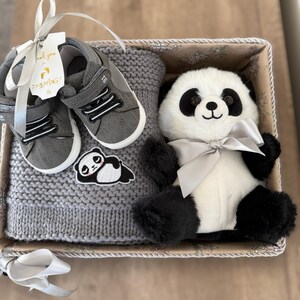 May include: A gift basket with a grey knitted hat, grey baby shoes, and a panda bear soft toy with a silver bow. The shoes have white soles and black details. A small panda patch is on the hat. A tag with text is attached.
