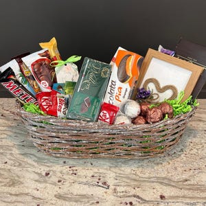 May include: A wicker basket overflowing with a selection of confectionery items, such as chocolate bars, biscuits, and sweets. The basket includes a framed picture with a heart design, and is lined with green decorative grass.