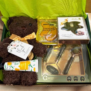 Peut inclure: Coffret cadeau garni d'un ours en peluche marron avec un nœud doré, d'un tube de lotion, de masques pour les yeux, d'un ensemble de rouleaux de jade et d'une boîte de chocolats. La boîte est doublée de papier de soie vert et contient une étiquette "Merci".