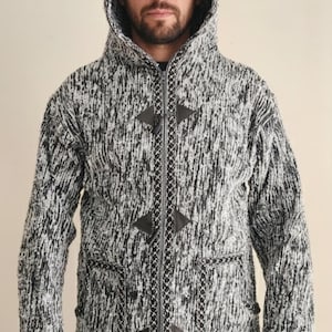 May include: A hooded knit jacket in black and white speckled yarn. The jacket features a toggle closure, two front pockets, and a decorative trim along the zipper and edges. The hood is pointed. The jacket is worn over a dark shirt.