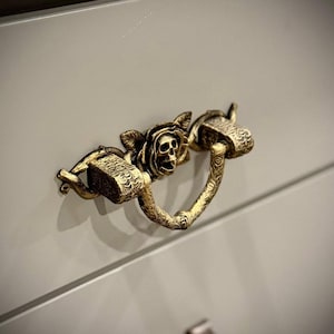 May include: A decorative brass drawer pull featuring a skull and floral design. The handle is attached to a light gray cabinet door. The skull is centered, with ornate details on the handle and surrounding elements.