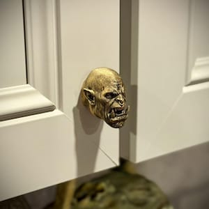 May include: A gold-colored cabinet knob in the shape of a grotesque orc face. The knob is mounted on a white cabinet door. The orc face has detailed features, including a snarling mouth and pointed ears. The cabinet door is part of a larger cabinet.