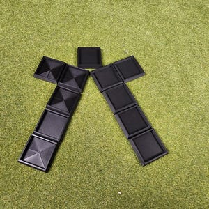 May include: Arrangement of black, square-shaped objects on a green surface. The objects are arranged in a stylized human figure. Some squares have textured surfaces, including pyramid and ribbed designs. The background is a solid green color.