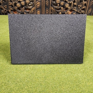 May include: A black, rectangular object with a textured surface. The object is positioned on a green surface, with a carved wooden background visible behind it. The object appears to be a box or container.