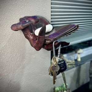 May include: A unique wall-mounted key holder shaped like a monster hand. The hand is a mix of dark red and purple with black claws, holding a brown leather wallet and a set of keys. A white stone rests on the hand.