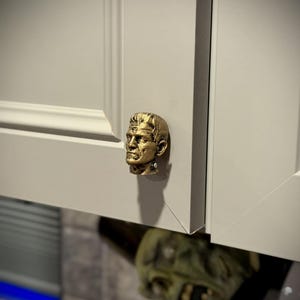 May include: A gold-colored cabinet knob in the shape of a sculpted head, mounted on a white cabinet door. The knob has detailed facial features. The cabinet door is closed. The overall theme is Halloween.