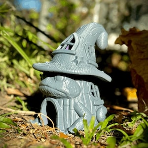 May include: A gray frog-shaped house figurine with a witch hat roof. The house has a door, windows, and a textured surface. The figurine is set outdoors among grass and leaves.