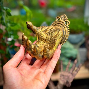 May include: A gold-colored wall-mounted hook shaped like a textured, clawed creature hand. The hand features sharp, pointed talons and a scaly skin texture, attached to a decorative, ornate circular base with a beaded edge and a fan-like top detail.
