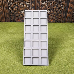 May include: A rectangular, light gray tray with a grid of twenty-four square compartments. The tray is made of a smooth, matte material and sits on a green surface. The background features a dark brown, carved wooden panel.