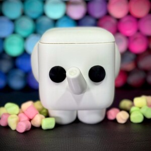 May include: A white, square-shaped elephant figurine with black eyes and a trunk. The figurine has rounded ears and is surrounded by colorful marshmallows. The background features a blurred array of colorful spheres.