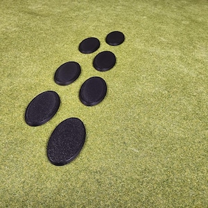 May include: A collection of seven black oval bases, likely for miniature figures, arranged on a green textured surface. The bases have a slightly rough texture and are uniformly black.