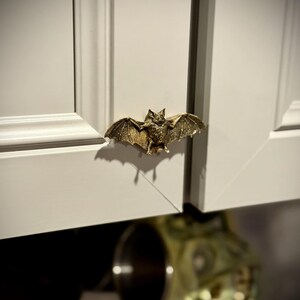 May include: A bronze-colored bat-shaped cabinet pull is mounted on a white cabinet door. The bat has outstretched wings and detailed features. The hardware adds a unique, gothic touch to the cabinetry.