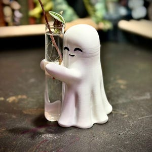 May include: A white 3D-printed ghost figurine with a smooth finish and a simple black-painted face, featuring two arms wrapped around a clear glass test tube filled with water and a small green plant cutting with variegated leaves.