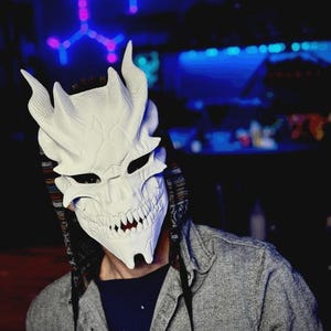 May include: A white, intricately designed mask with sharp teeth and horns. The mask is worn with a gray jacket and a dark blue shirt. The background features a neon light display with a blue and pink color scheme.