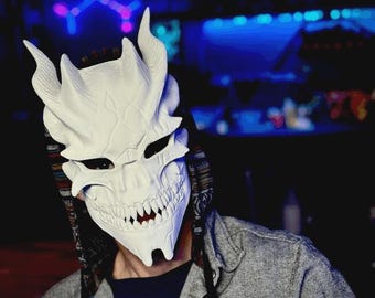 May include: A white, textured, full-face mask featuring sharp, horn-like protrusions at the top, narrow eye slits, and a prominent, jagged, toothy grin. The mask is worn over a dark shirt and a grey button-down shirt with a patterned hood lining.