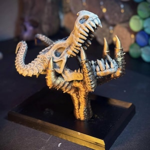 May include: A detailed, gold-colored dragon skull sculpture with sharp teeth and horns, mounted on a black rectangular base. The sculpture has a textured appearance and is likely a decorative item or collectible.
